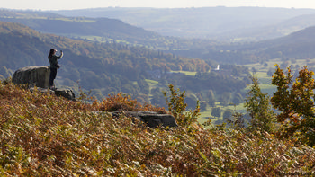 Curbar Edge photographer This landscape photograph shows a photographer standing on Curbar Edge, capturing the sweeping rural scenery of the Peak District in Derbyshire, United Kingdom. Taken during the early afternoon in autumn, the image features vibrant brown and orange foliage typical of the season, while the gentle sunlight highlights the layered hills and valleys below. The scene showcases the natural beauty of the Peak District, with lush greenery, patches of forest, and open countryside stretching into the distance, characteristic of this well-known region.